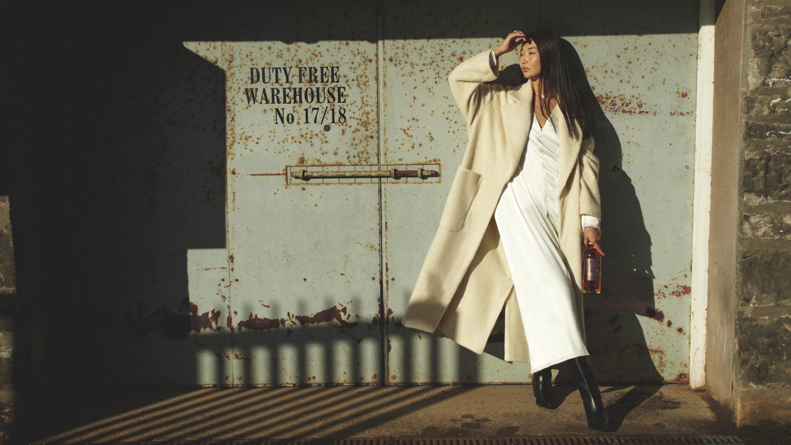 A woman standing in front of a warehouse door holding a bottle of 22 Year Old Longmorn Whisky
